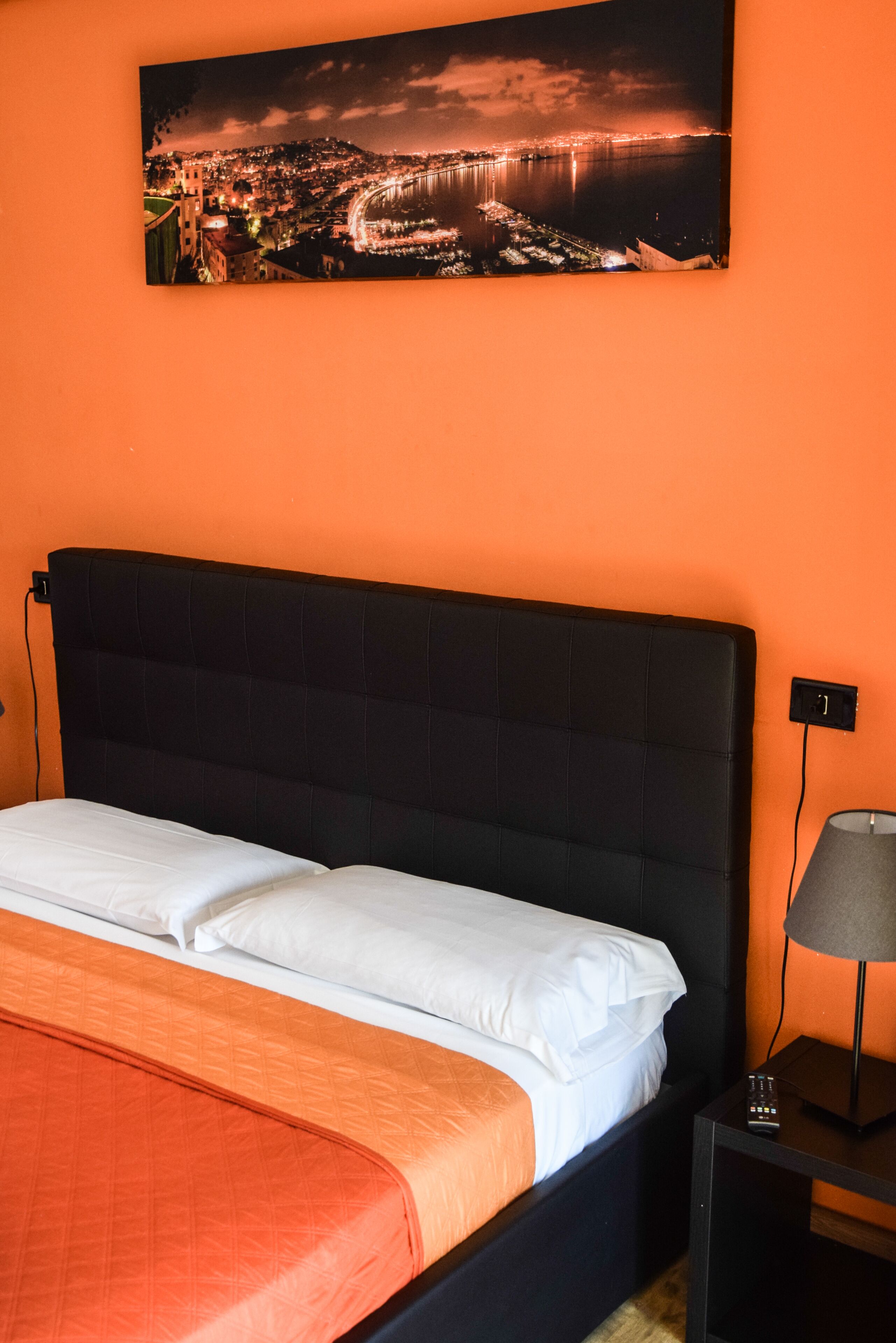 Una cama con cabecera negra, ropa de cama blanca y una manta naranja. Una mesita de noche con lámpara y control remoto está al lado de la cama.