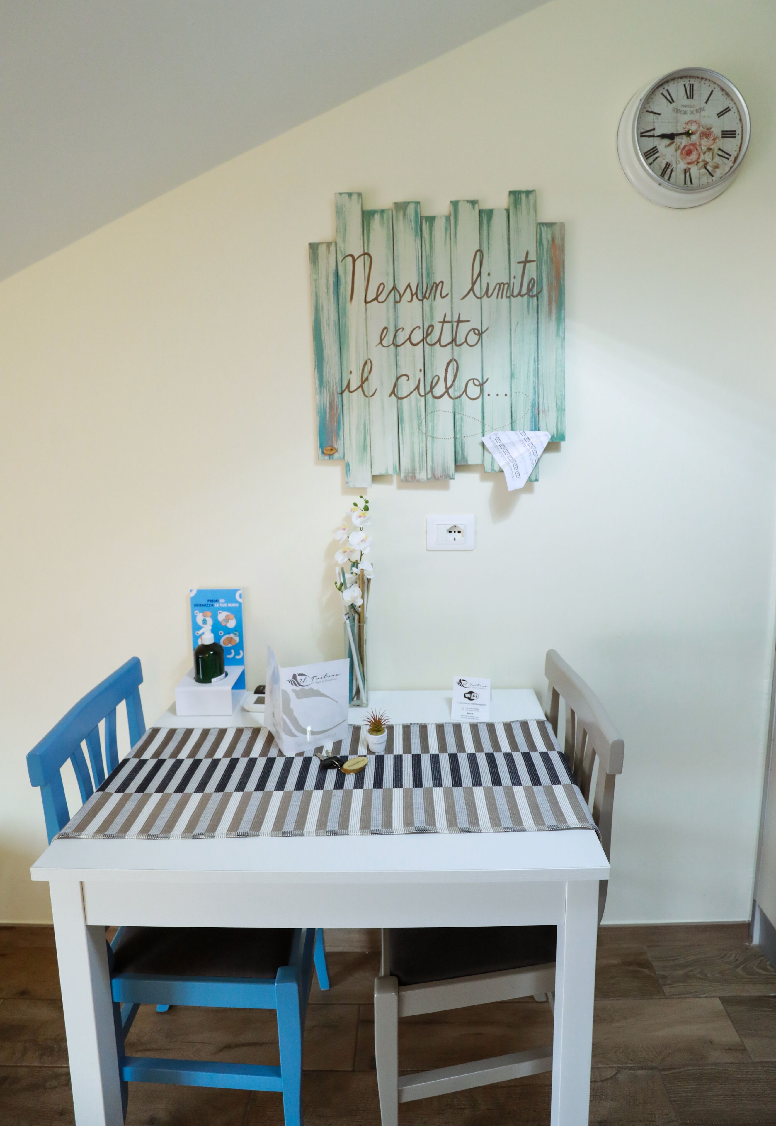 Un comedor con una mesa blanca y sillas azules, un adorno en la pared, un reloj y un jarrón con flores.
