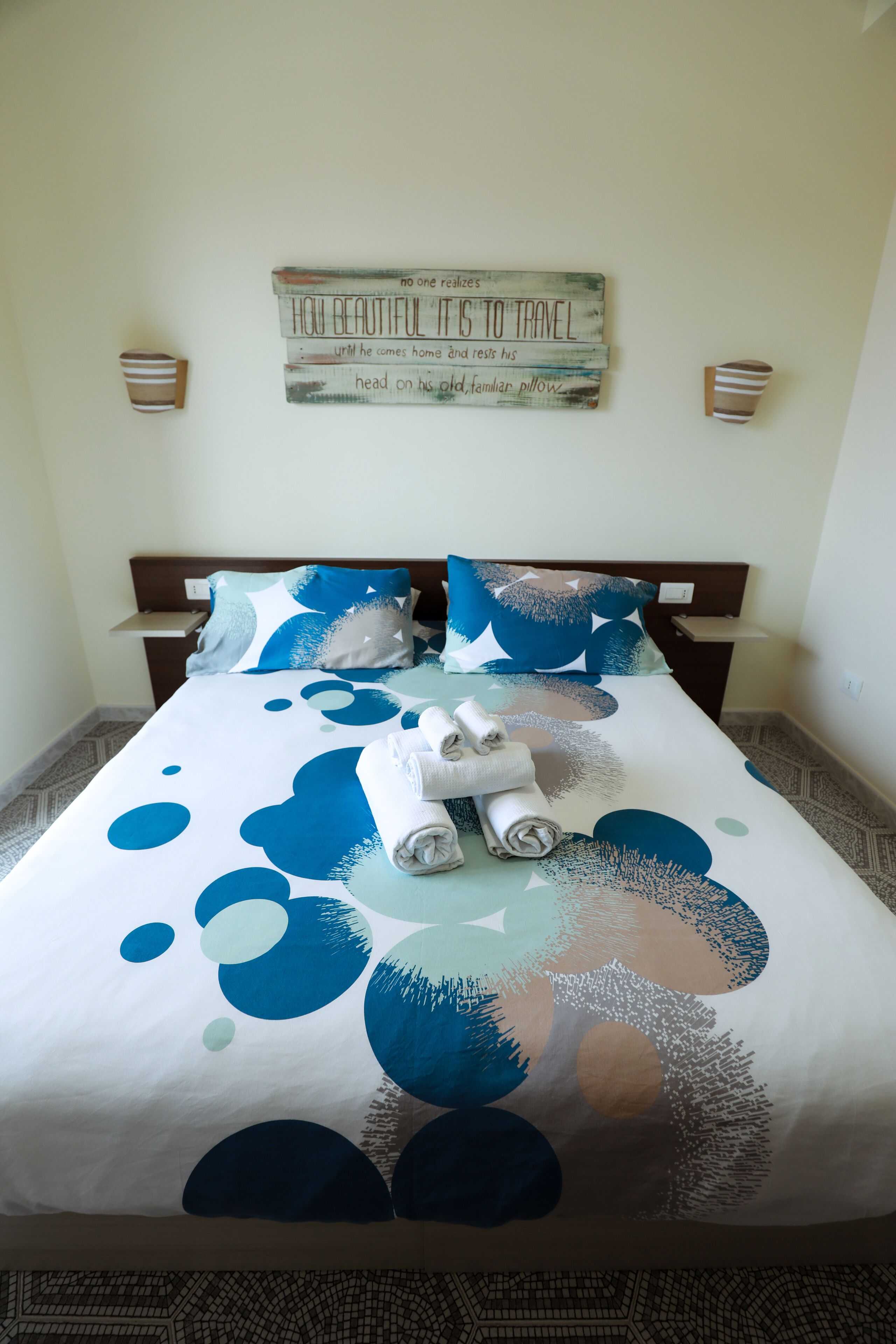 Una cama bien hecha con un estampado abstracto azul y blanco, un cojín con diseño similar y un letrero colgado en la pared con texto.
