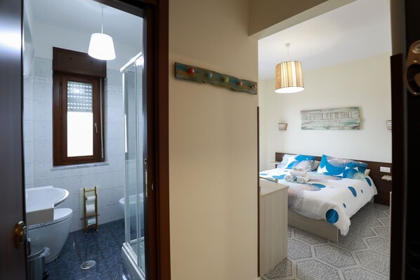 Un baño con inodoro, lavabo y ducha. Hay una cama con colcha y almohadas azules y blancas.