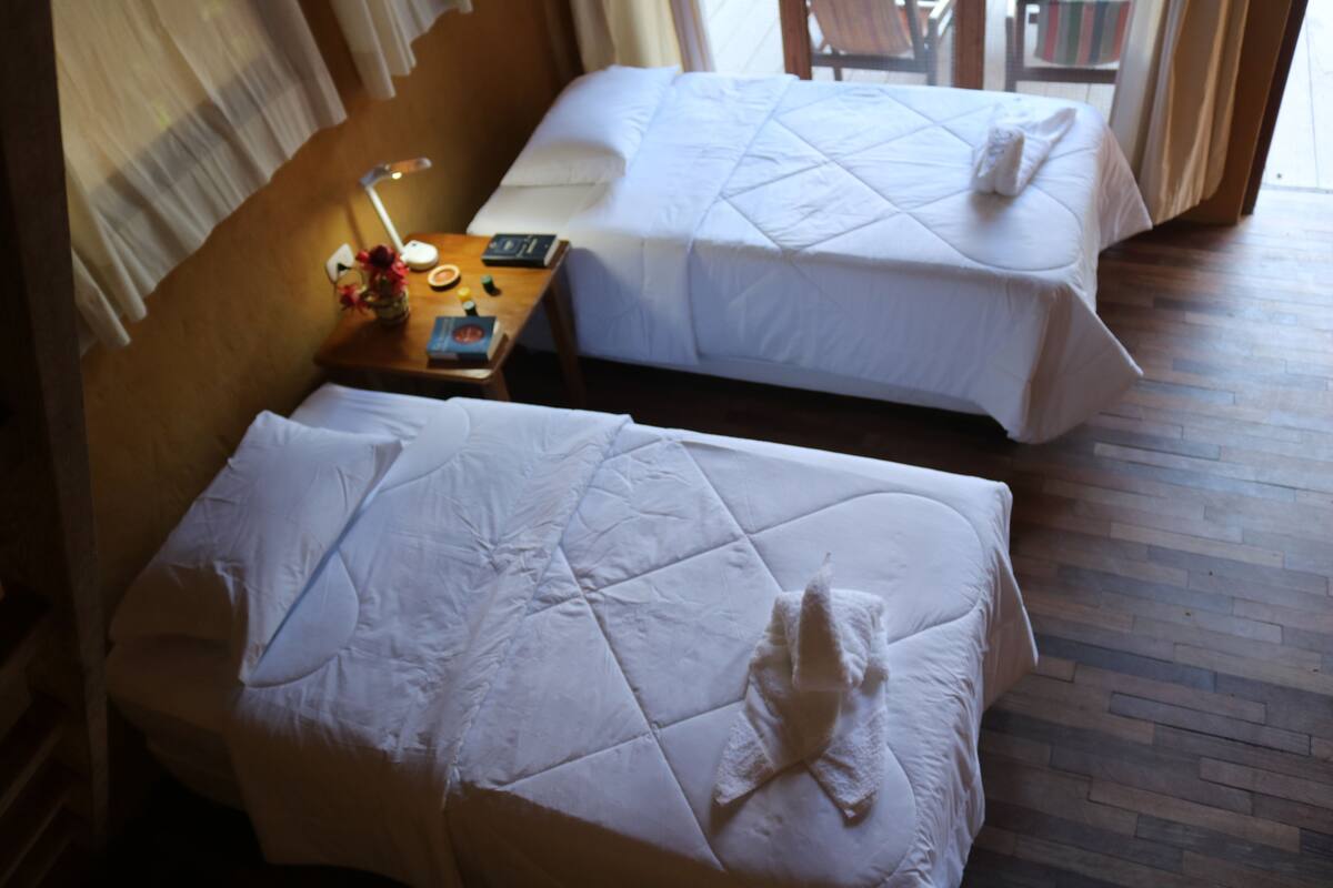 A hotel room with two beds, wooden flooring, and a small table with a lamp and a book.