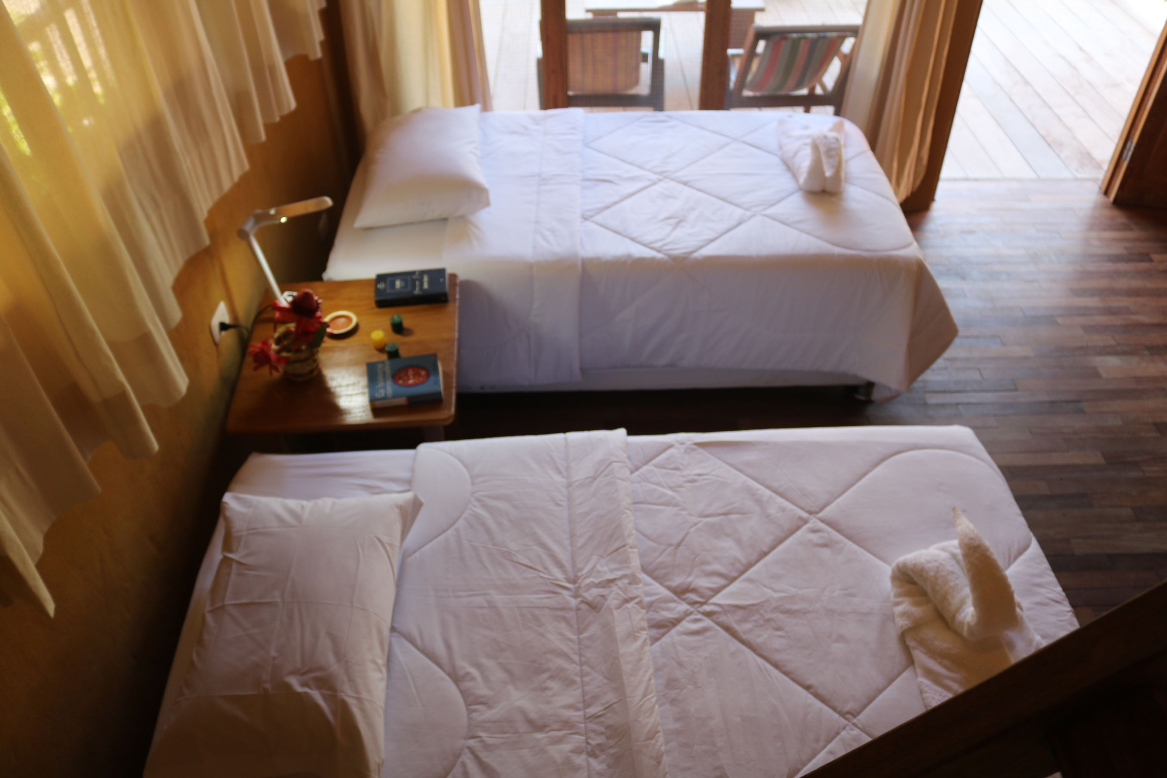A hotel room with two beds, a wooden floor, a nightstand with a lamp, and a window with curtains.