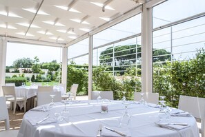 Un espace extérieur abrité, aménagé pour les repas, avec des tables rondes dressées, et entouré de grandes fenêtres donnant sur un jardin luxuriant.