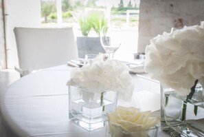 Une table surmontée de vases en verre transparent contenant des fleurs blanches, une chaise blanche et une fenêtre donnant sur un paysage verdoyant.