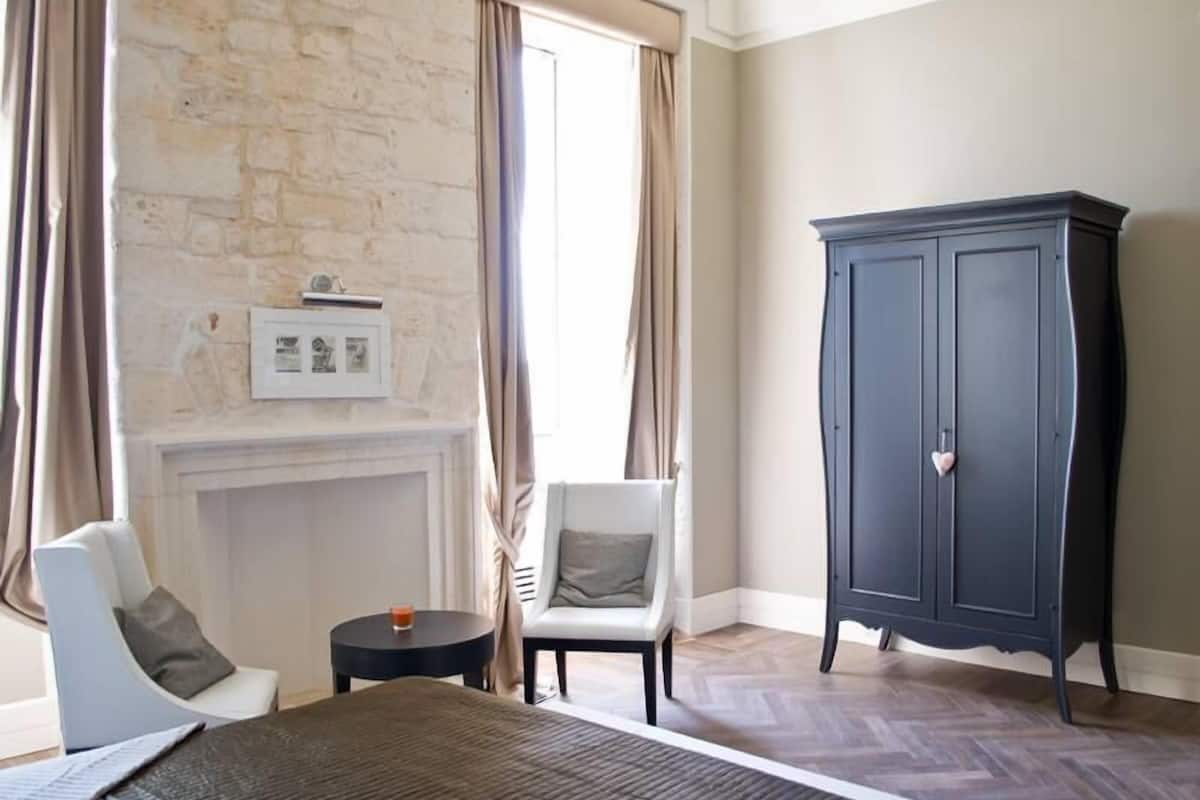 A bedroom with a bed, a chair, a small table, a cabinet, and a fireplace.