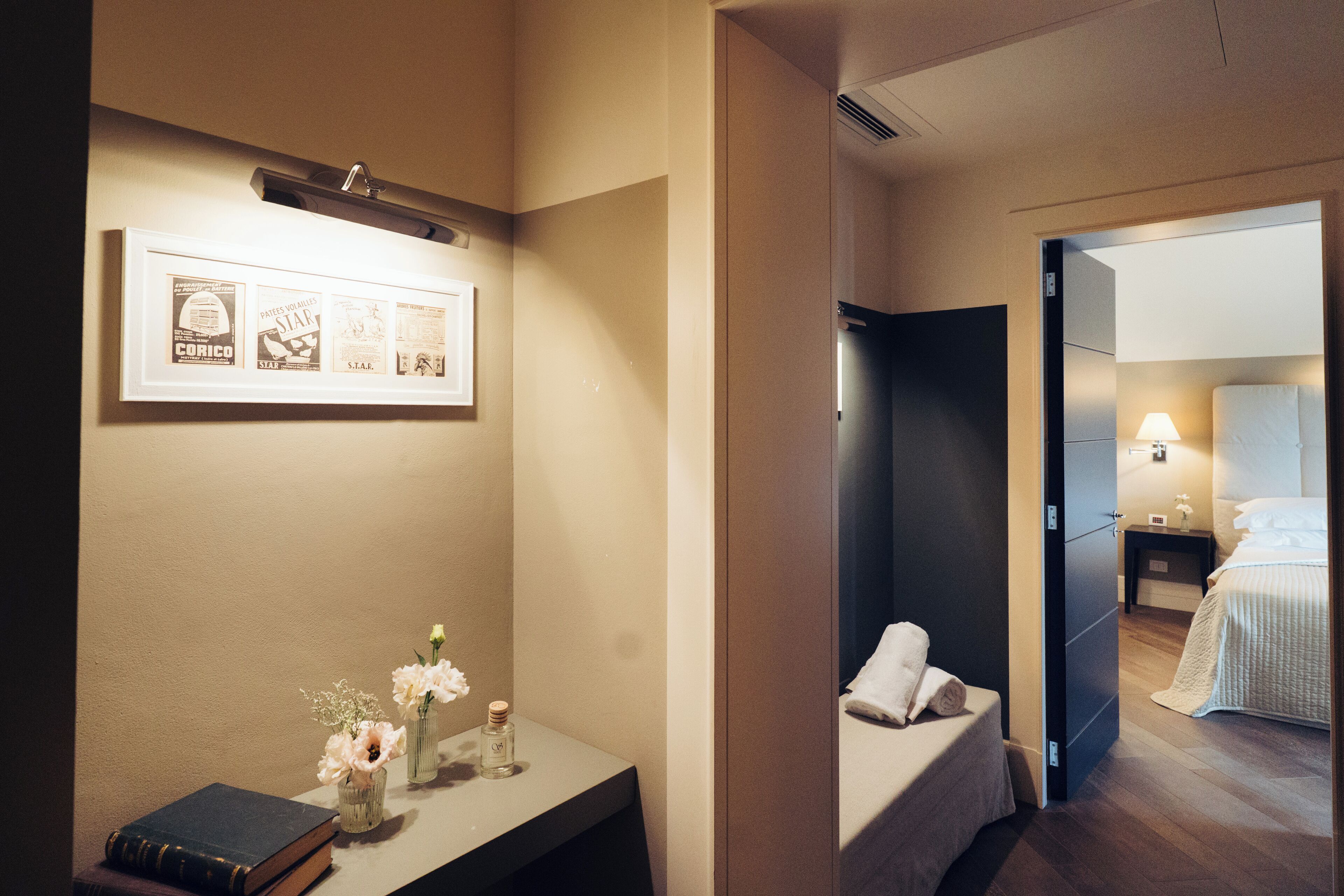 A hotel room with a bed, a bedside table with a book and flowers, a framed picture on the wall, and a nightstand with a lamp.