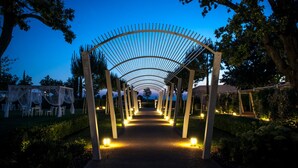 A pathway under a white, arched structure with evenly spaced columns, surrounded by trees and outdoor seating.
