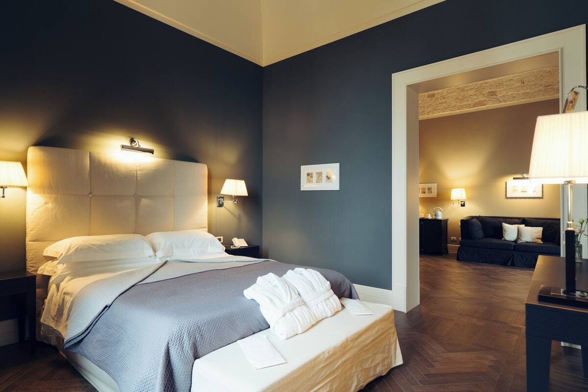 A modern hotel room with a large bed, bedside lamps, a dark wall, and a wooden floor.