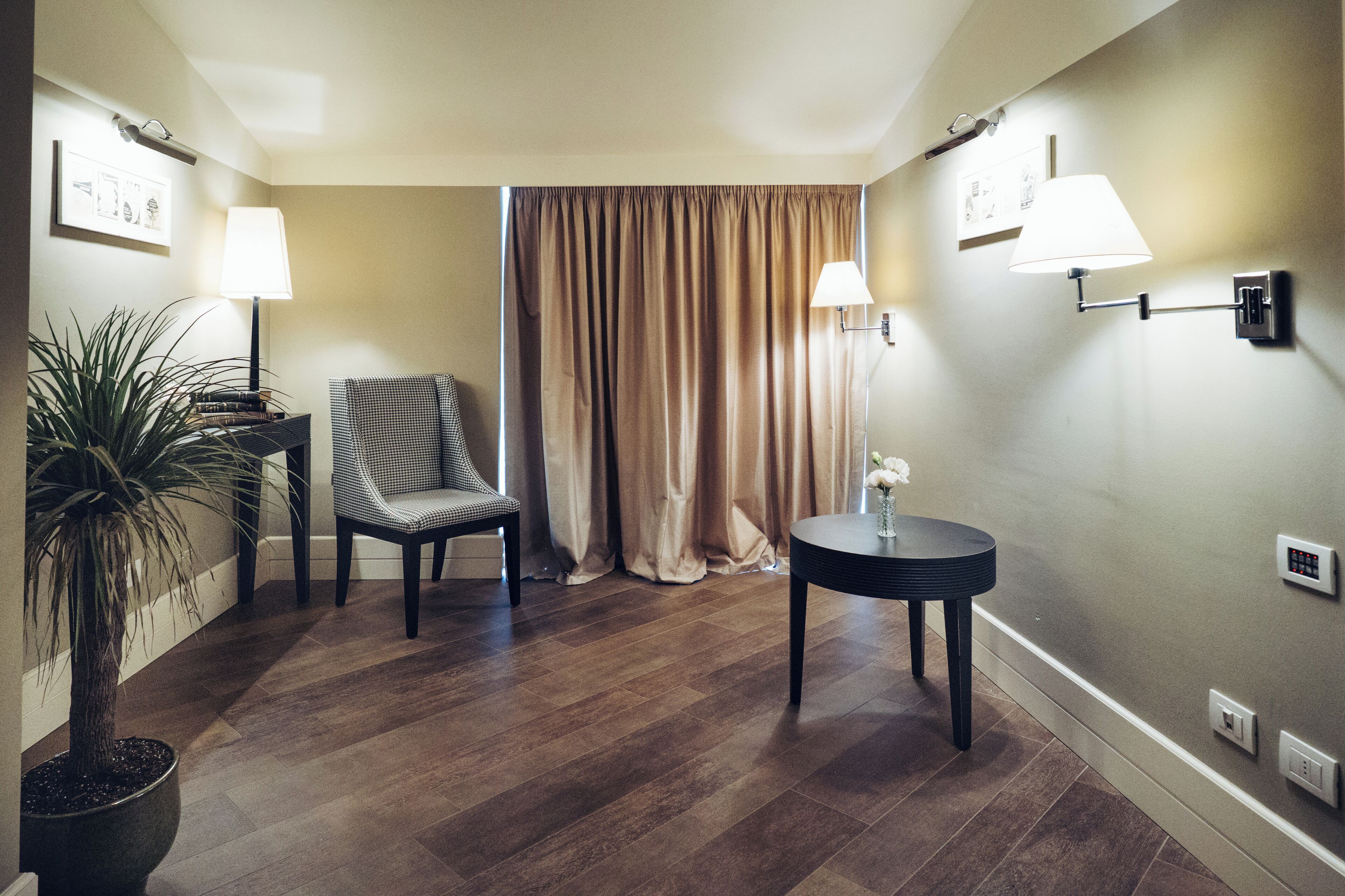 A hotel room with a wooden floor, a chair, a table, a potted plant, and two wall-mounted lamps.