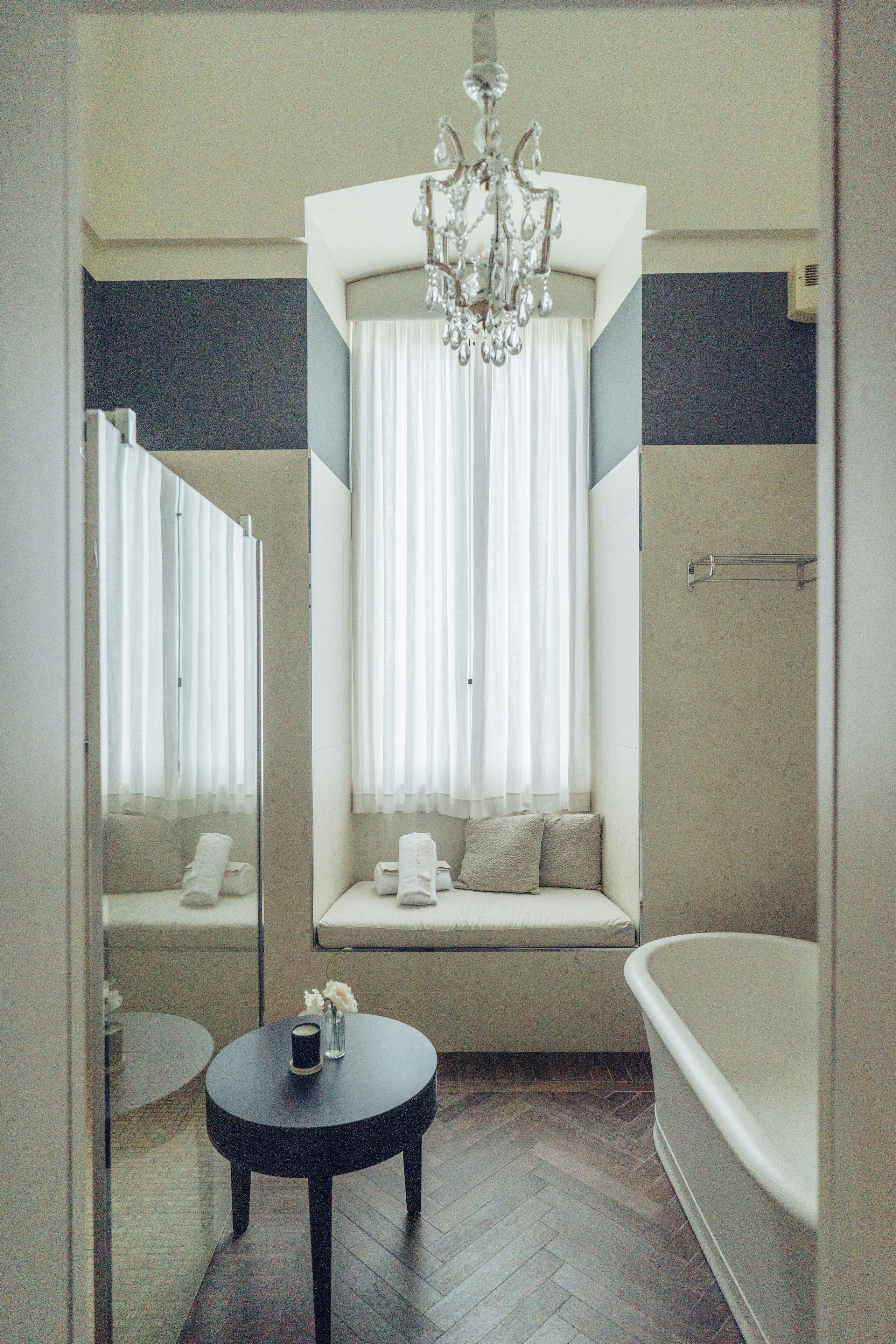 A bathroom with a bathtub, a window seat with curtains, a round table with a candle, and a chandelier.