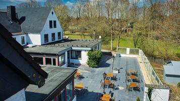 En tagterrasse med udendørs møbler, en moderne bygning med et skråt tag, og udsigt over et landskab med træer.