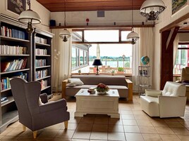 Ein Wohnzimmer mit einer Couch, Sesseln, einem Couchtisch und einem Bücherregal. Ein Fenster bietet Blick auf ein Gewässer und Grünflächen.