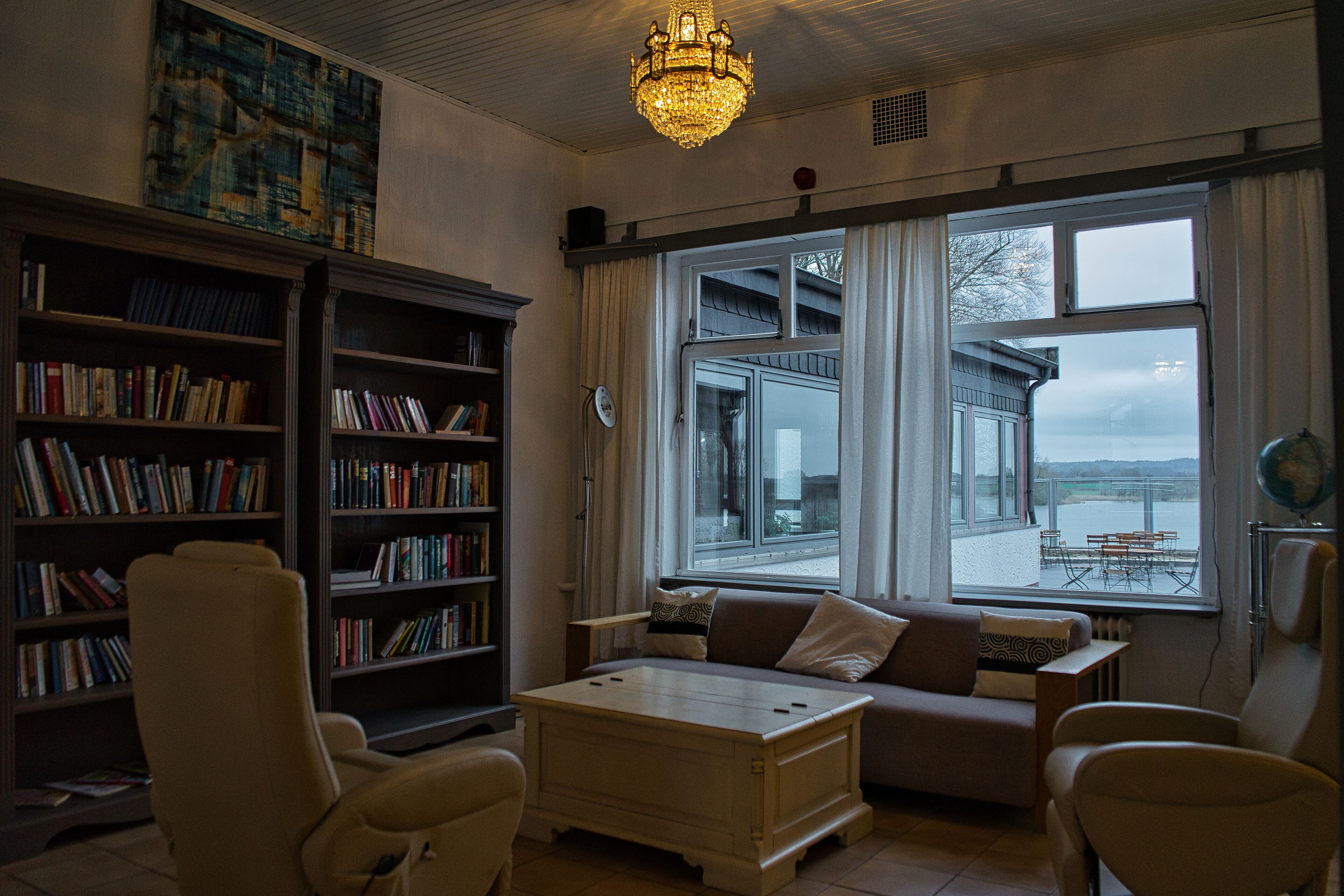Ein gemütliches Wohnzimmer mit einer Couch, Sesseln, einem Couchtisch und einem Bücherregal voller Bücher. Ein großes Fenster bietet Blick auf einen Balkon und ein Gebäude gegenüber dem Wasser.