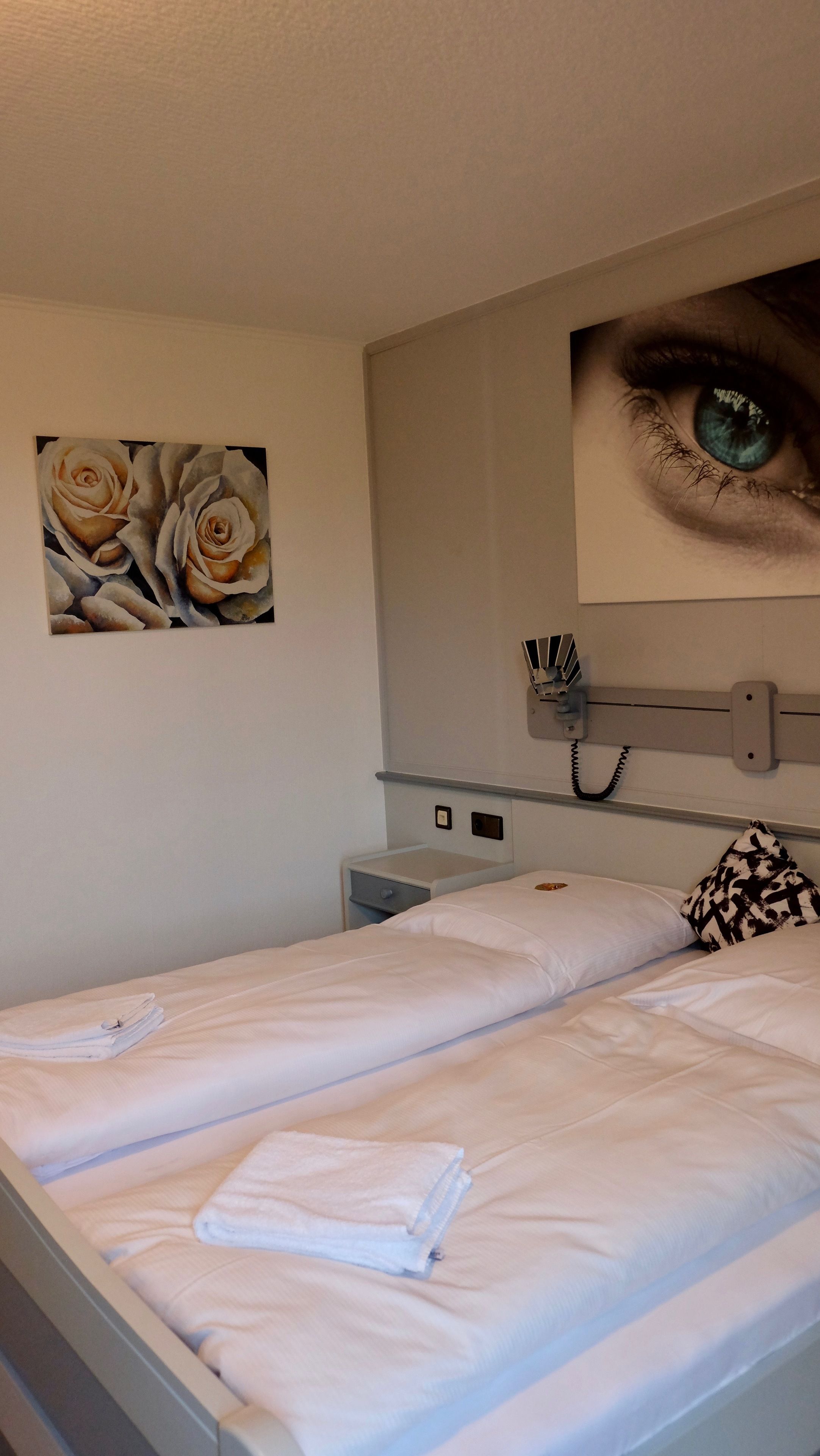 A bedroom with two beds, a painting of roses, a large eye artwork, and a bedside table with a lamp.