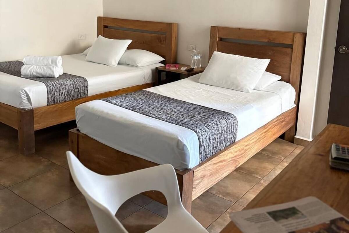 Una habitación de hotel con dos camas de madera, una silla blanca, una mesita y un cuadro en la pared.