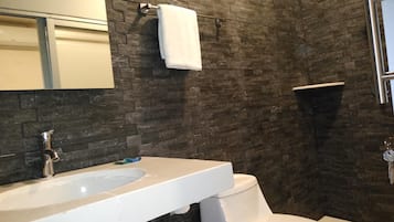 Un baño con un lavabo blanco, un inodoro y un espejo. Las paredes están cubiertas de baldosas de piedra oscura.