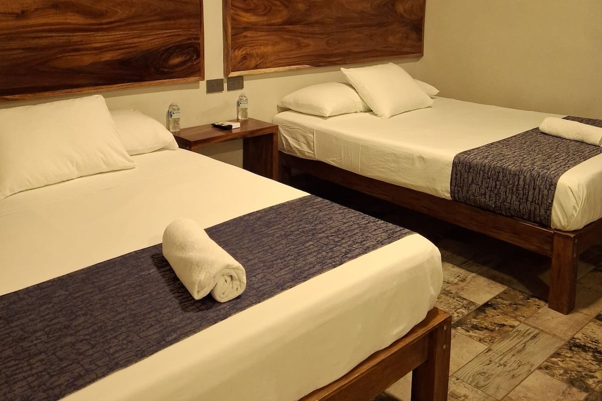 Habitación de hotel con dos camas, cabeceros de madera, ropa de cama blanca y una mesita de noche con una botella y un control remoto.