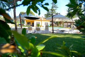 A poolside area with lounge chairs and umbrellas, surrounded by greenery and a building with a yellow facade. - B&B il giardino di Agata (Mascalucia)