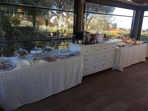 A buffet setup with various food items and a coffee station, set against a backdrop of a large window with a view of trees and a building. - B&B il giardino di Agata (Mascalucia)