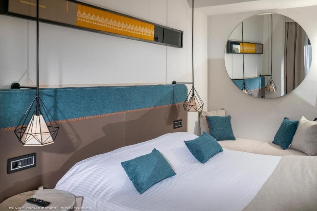 Quarto de hotel moderno com uma cama grande, um espelho redondo e luminárias pendentes.