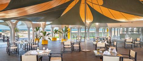 An outdoor dining area with tables and chairs, large umbrellas, and a view of the beach.