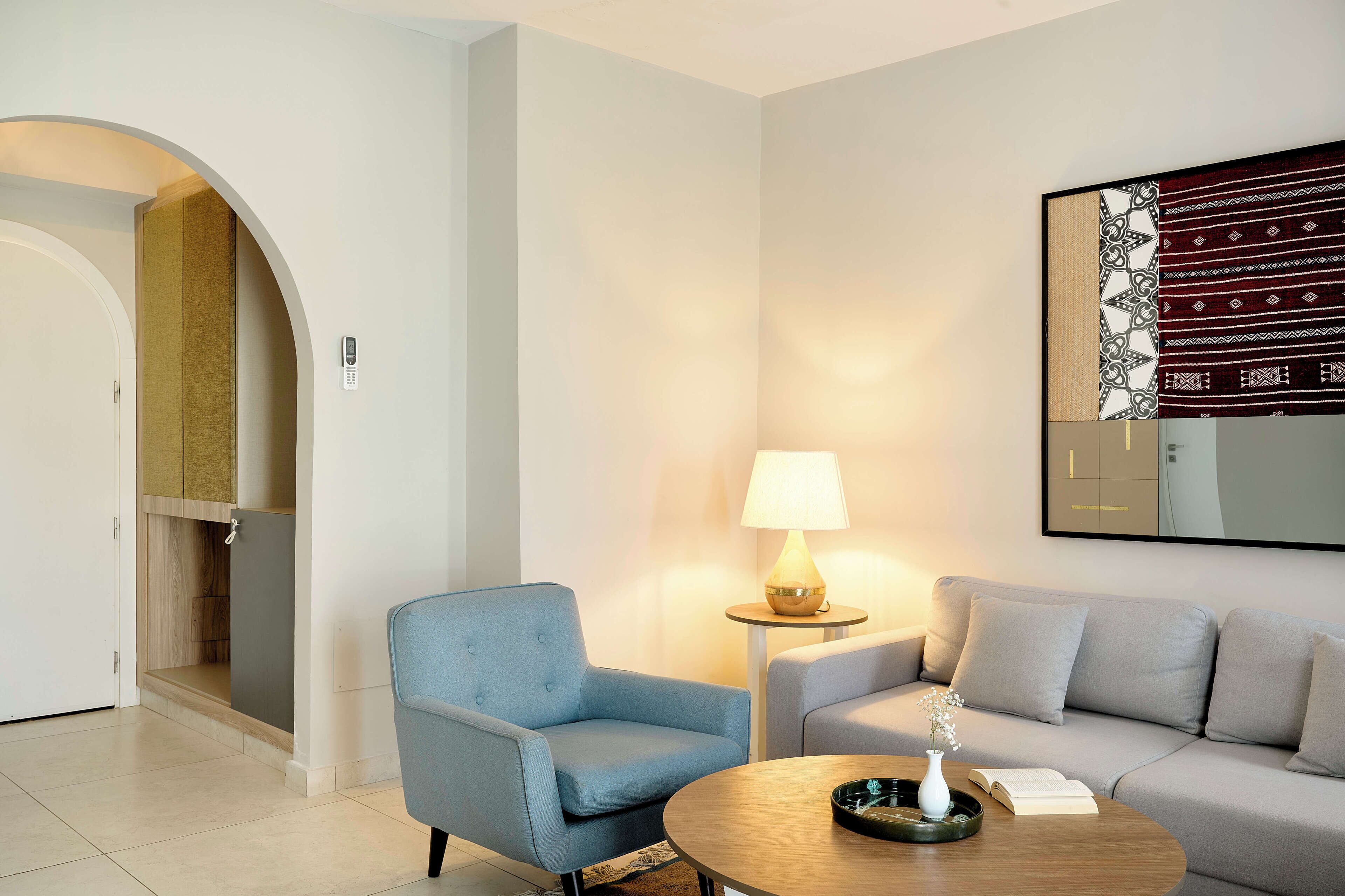 Habitación de hotel moderna con un sillón azul, un sofá gris, una mesa redonda con un libro y un florero, y una lámpara sobre una mesita auxiliar.
