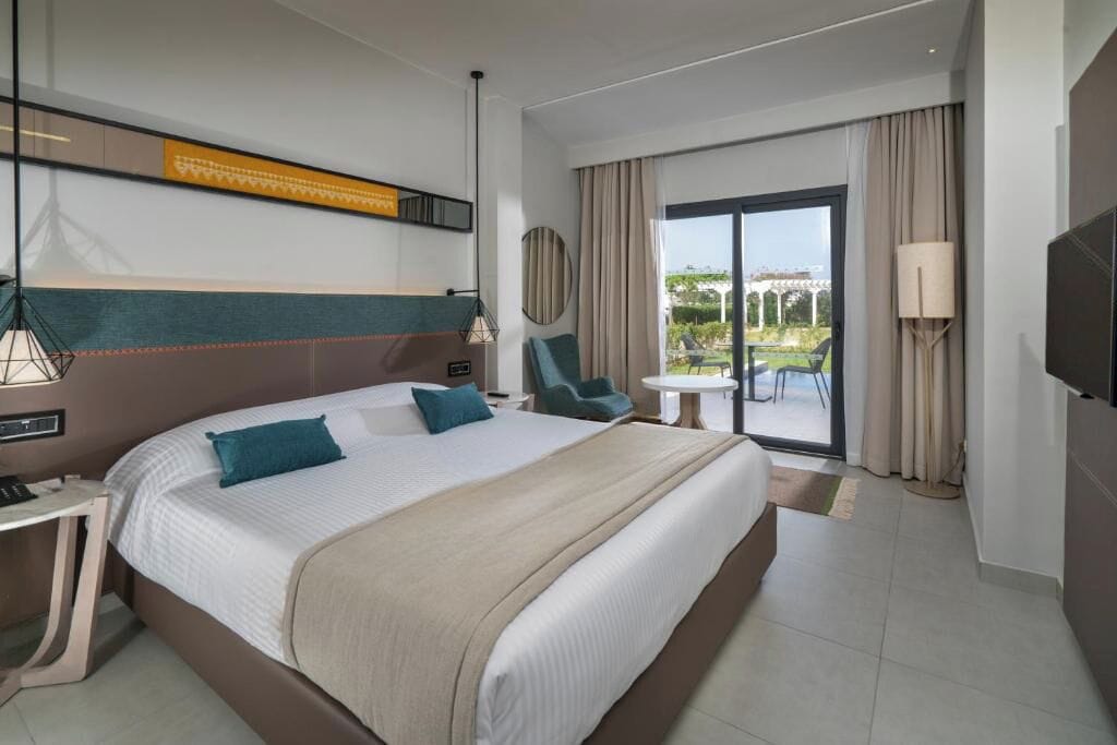 A hotel room with a large bed, a desk, a chair, a TV, and a balcony with a view.