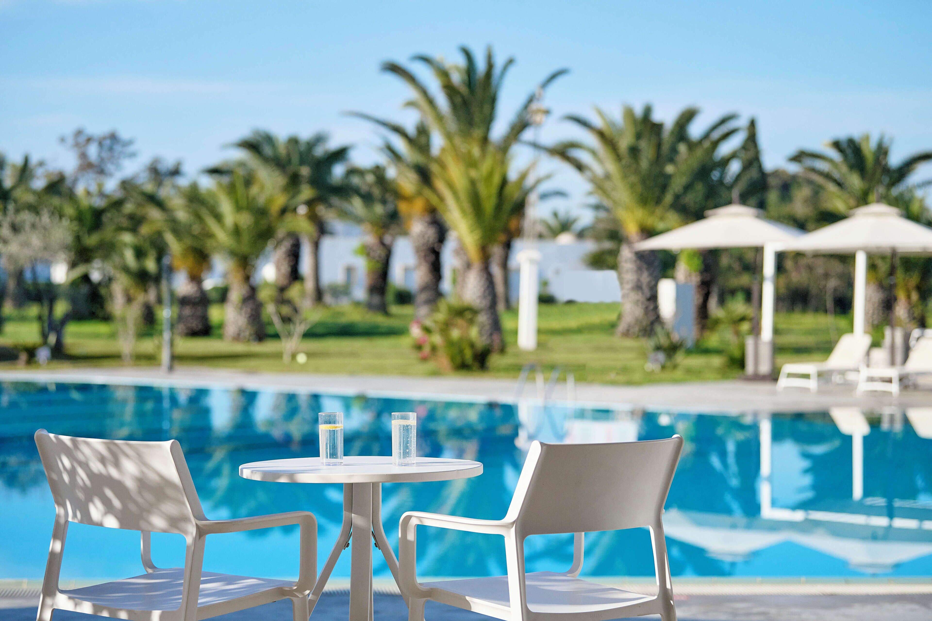 A poolside area with two white chairs and a table, surrounded by palm trees and a clear blue sky.