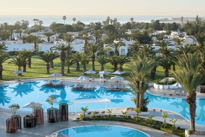 Zona de piscina con sillones de descanso y sombrillas, rodeada de palmeras y edificios blancos.