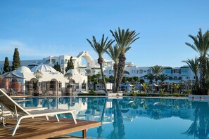 A pool area with lounge chairs, palm trees, and a white building with arches in the background. - The Mirage Resort & SPA (Hammamet)