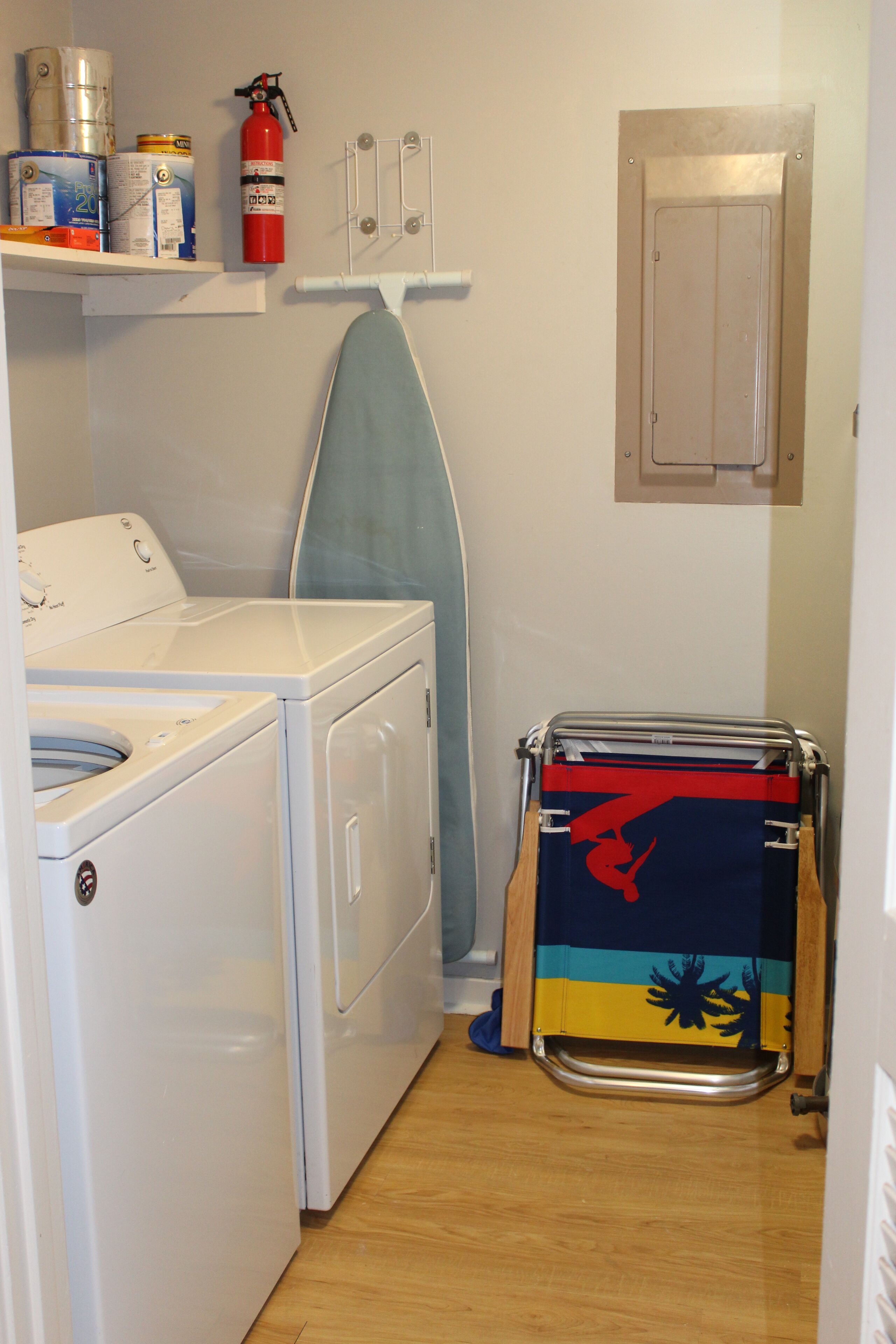 A laundry area with a washing machine, dryer, ironing board, and a fire extinguisher on a shelf.