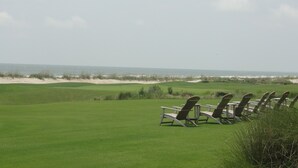 A golf course with chairs and tables arranged for guests, a clear view of the ocean, and sand dunes in the background.
