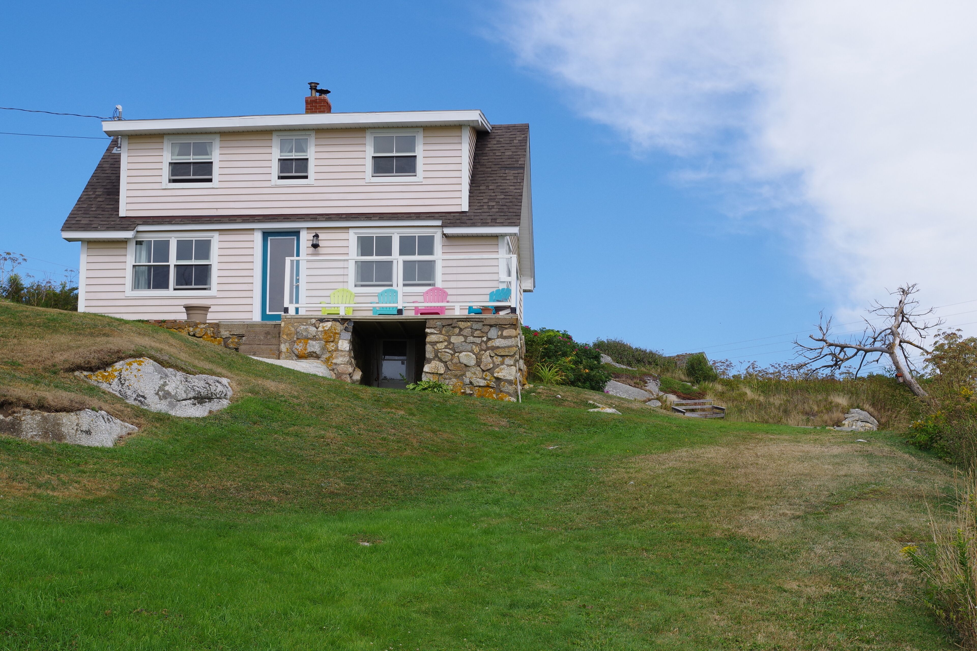 Une maison à deux étages avec une entrée en pierre, une véranda agrémentée de chaises Adirondack aux couleurs vives et une colline verdoyante qui entoure la propriété.