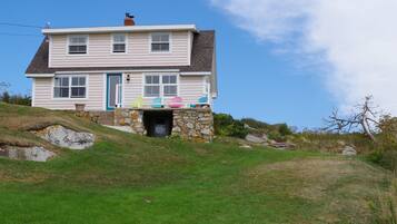 Une maison à deux étages avec une entrée en pierre, une véranda agrémentée de chaises Adirondack aux couleurs vives et une colline verdoyante qui entoure la propriété.