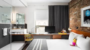 A modern hotel room with a glass shower, a large bed, a desk with a computer, and a brick wall.