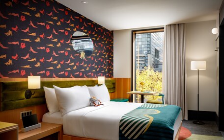 A hotel room with a large bed, a nightstand, a desk, and a view of the city through a large window.