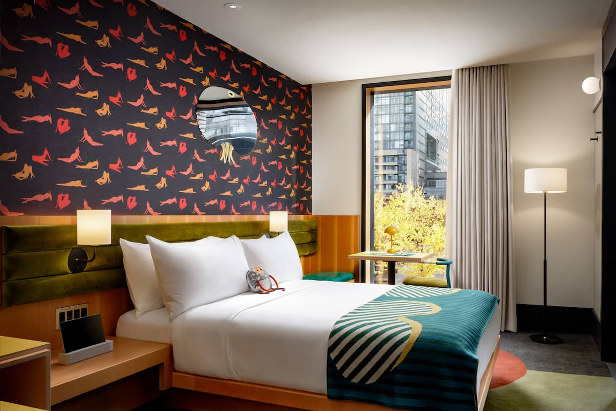 Habitación de hotel con una cama grande, una mesita de noche, un escritorio y vistas de la ciudad a través de un ventanal amplio.