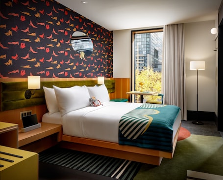 A hotel room with a large bed, a nightstand, a desk, and a view of the city through a large window.