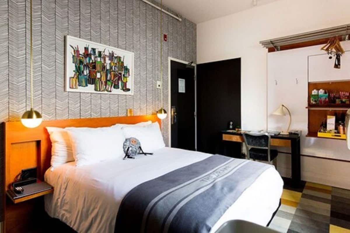 Habitación de hotel con cama, escritorio y silla. Hay un cuadro en la pared y una estantería con objetos.