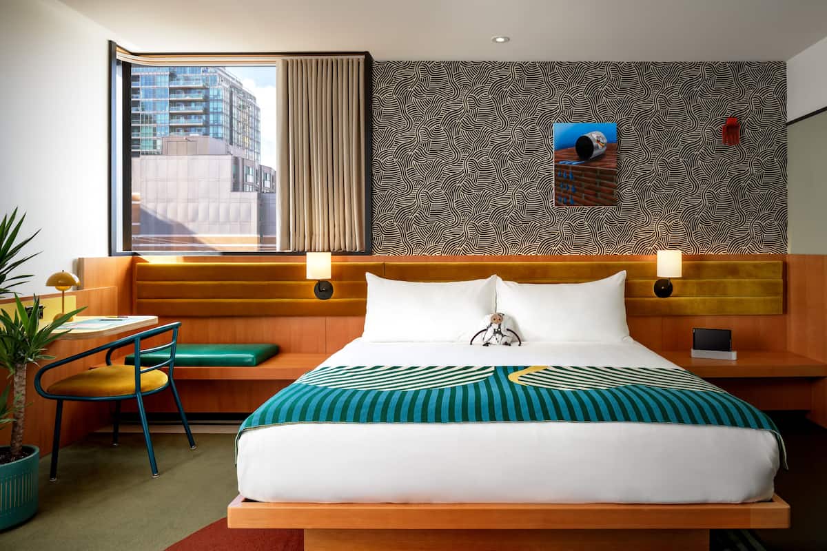 Habitación de hotel moderna con una cama grande, un escritorio con una silla, una planta en maceta y vistas a un paisaje urbano a través de la ventana.