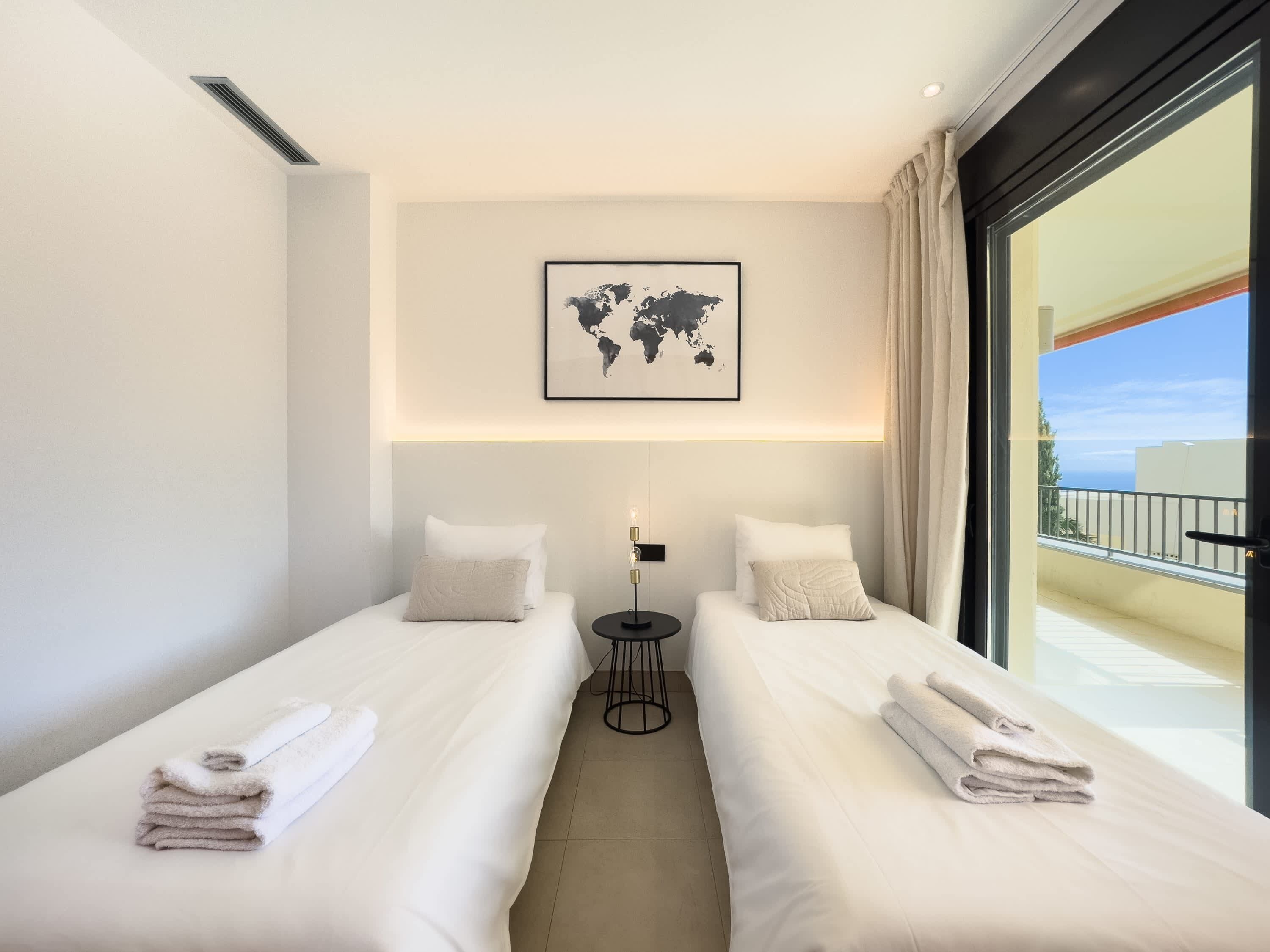 Habitación de hotel con dos camas, un mapa mundial en la pared, una mesita y vista al mar.