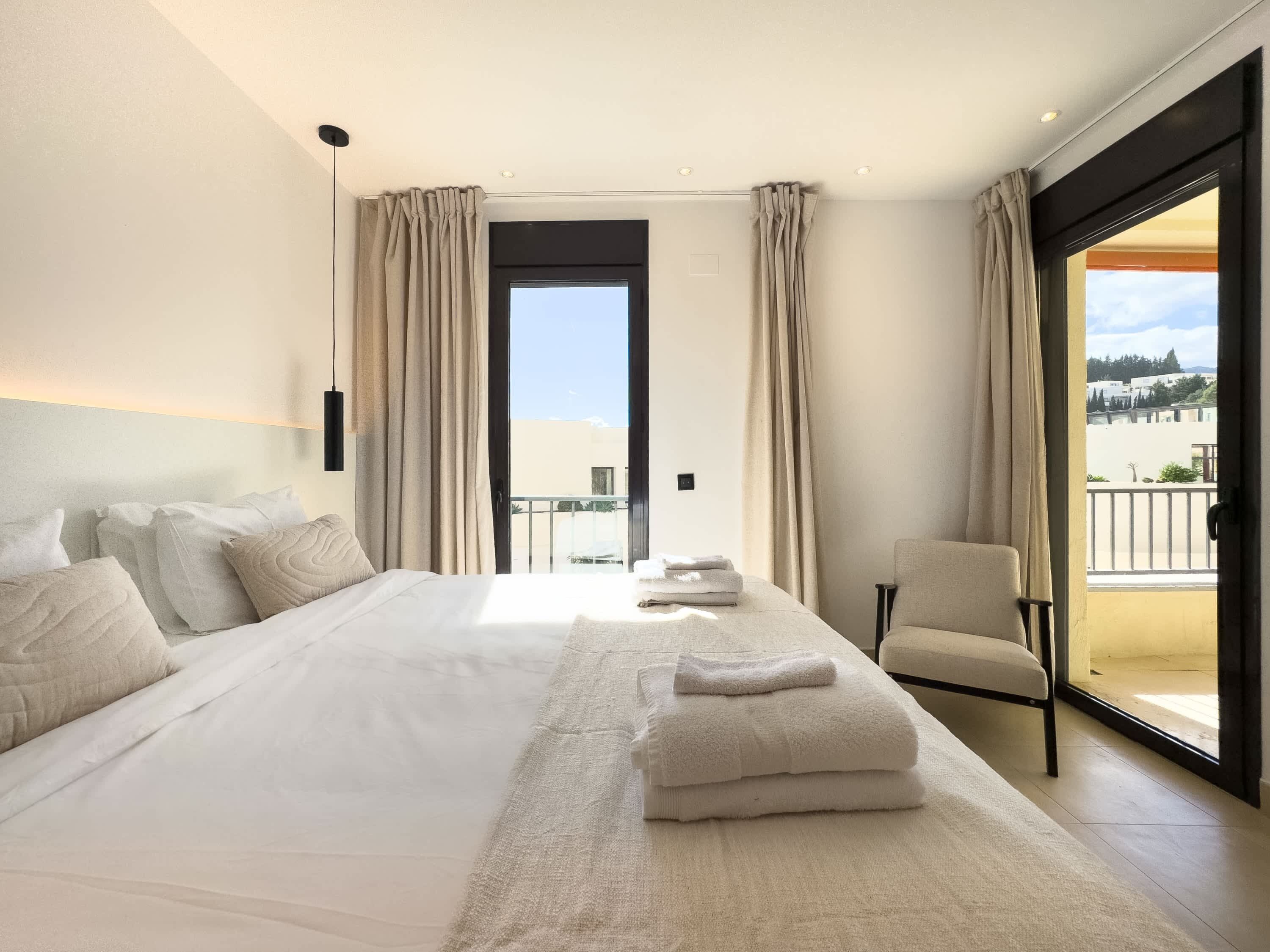 Uma cama grande com roupa branca, uma cadeira e vista para o exterior através de uma porta.