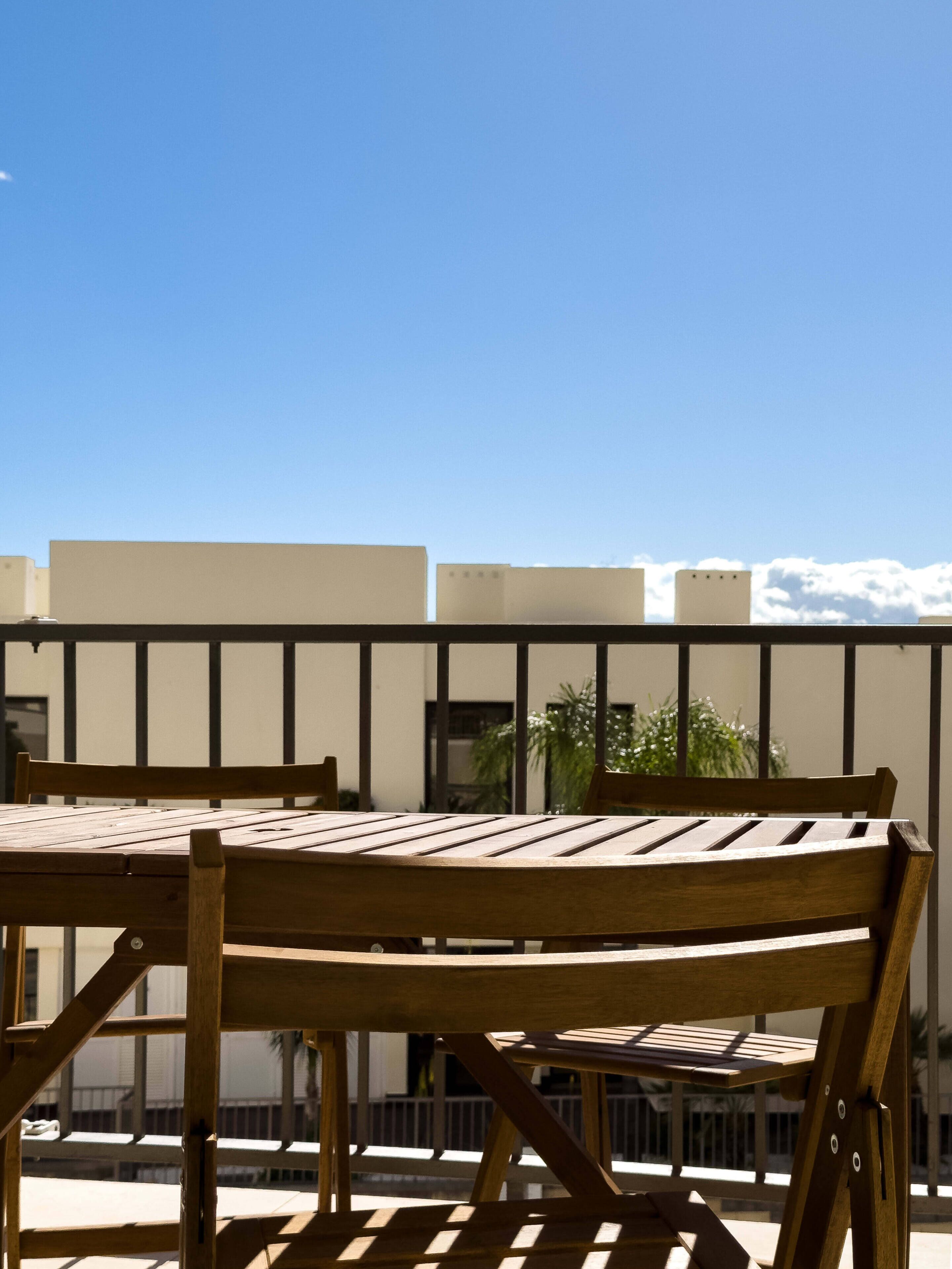 A wooden table and chairs on a balcony with a clear sky and buildings in the background.