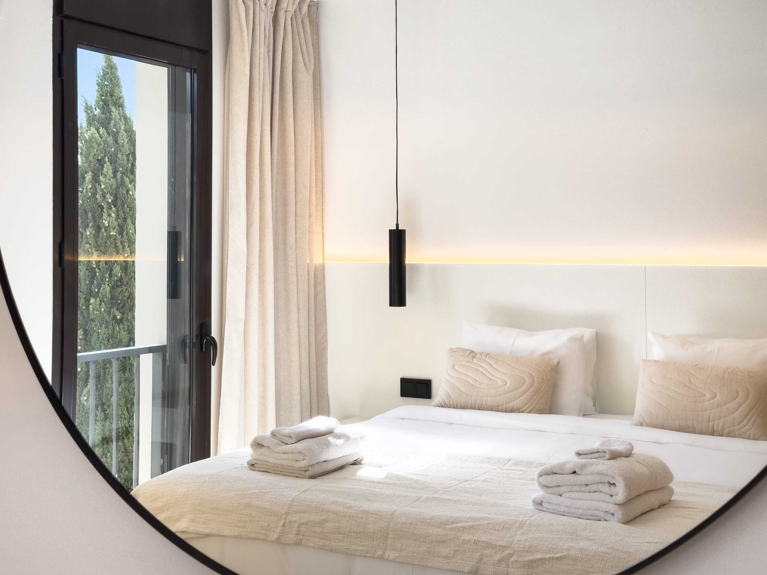 A modern bedroom with a large bed, two pillows, a nightstand, and a view of trees through a window.