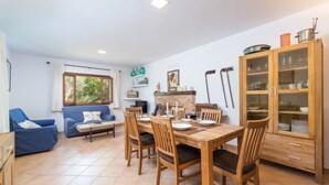 Ein Essbereich mit einem Holztisch, gedeckt für vier Personen, umgeben von Holzstühlen. Rechts befindet sich eine blaue Couch und ein Holzschrank mit Glastüren.