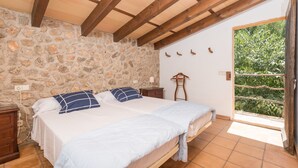 Ein Schlafzimmer mit zwei Betten, einer Steinwand, Holzbalken, einem Nachttisch, einer Lampe und Blick auf Grün hinter einer offenen Tür.