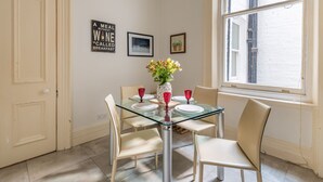 A dining area with a glass table, four chairs, a vase with flowers, and a window with a view.