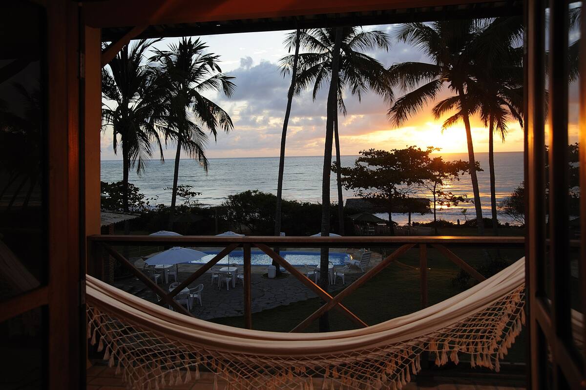 A hammock overlooking a pool area with palm trees and a sunset view.