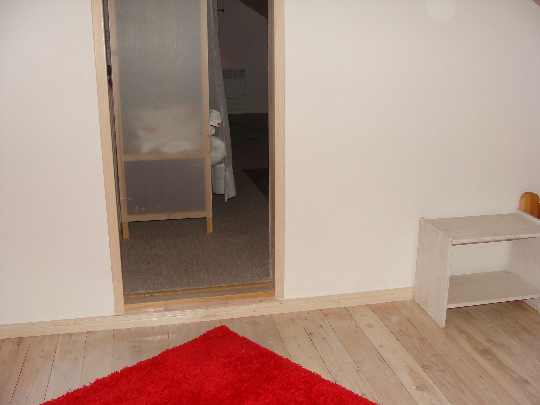 A room with a wooden floor, a red rug, a white wall, and a small white table.