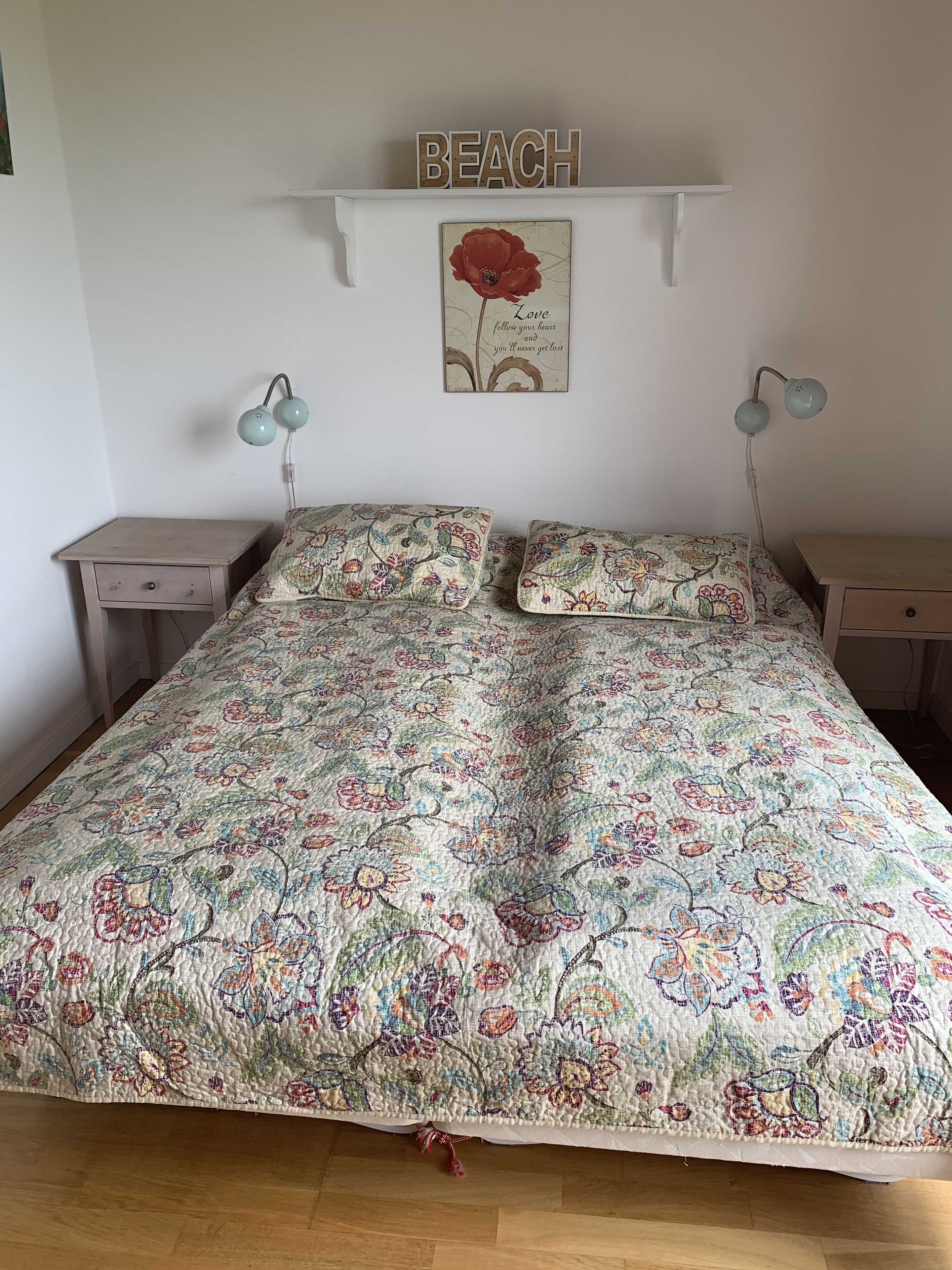 A bedroom with a floral bedspread, a 'BEACH' sign, a framed quote, and two bedside lamps.